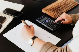 pexels-karolina-grabowska-4476375 Top-down view of a professional accountant using a calculator and writing notes on financial ledger paperwork at a dark desk with a wooden keyboard and smartphones nearby.